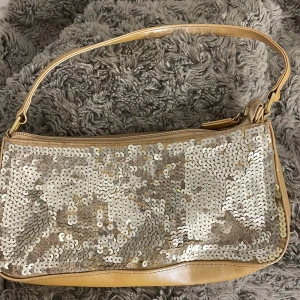 Guld axelväska med paljetter - Snygg guld axelväska med glittriga paljetter på framsidan. Perfekt för att lägga till lite bling till din outfit. Väskan har en praktisk dragkedja upptill och en bekväm axelrem.