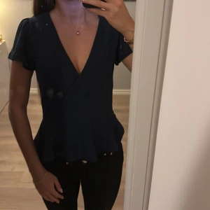 Mörkblå omlottblus från Holly & White - Säljer en elegant mörkblå omlottblus från Holly & White. Blusen har korta ärmar och en v-ringad design som ger en stilren look. Perfekt för att klä upp eller ner beroende på tillfälle.