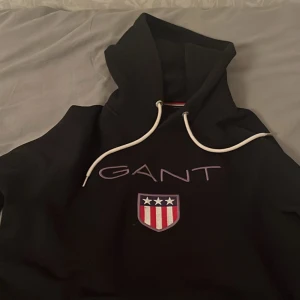 Svart hoodie från GANT - Snygg svart hoodie från GANT med vit dragsko och ett broderat emblem på bröstet. Perfekt för en avslappnad stil. Den har en klassisk passform och är tillverkad i mjukt material för extra komfort.