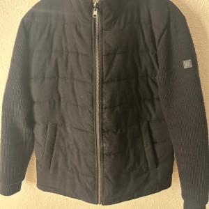 Brunn & stengade cardigan - Snygg svart cardigan jacka från Brunn & stengade dragkedja och långa ärmar. Jackan har en stilren design med ribbade ärmar och en diskret logga på ena ärmen. Perfekt för kyligare dagar. 