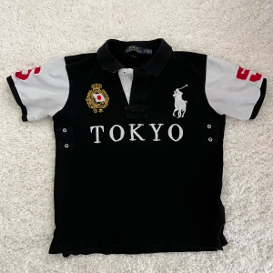 Svart pikétröja från Ralph Lauren - HELT NYA RALPH LAUREN POLO T SHIRT. Snygg svart pikétröja från Ralph Lauren med texten 'TOKYO' på framsidan. Tröjan har vita detaljer och ett broderat emblem på bröstet. Kortärmad design med klassisk krage och knappar.