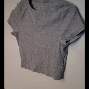 Grå ribbad magtröja  - Säljer en enkel och stilren grå ribbad t-shirt med korta ärmar. Perfekt för en avslappnad look. T-shirten har en rund halsringning och är i en mjuk och bekväm kvalitet.