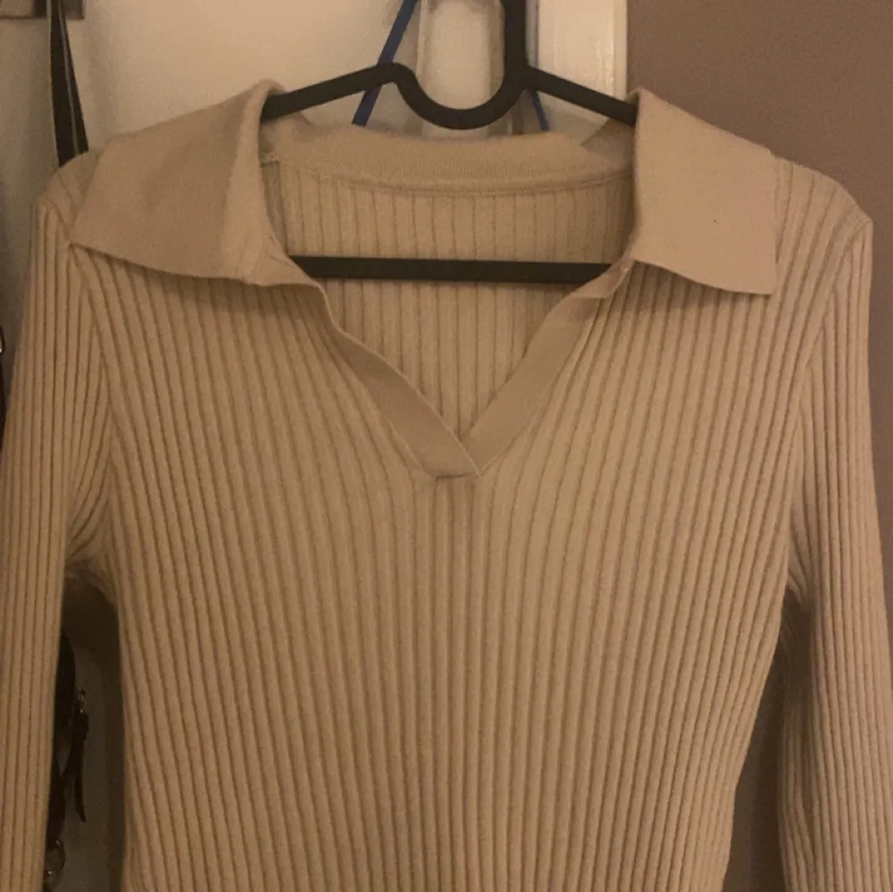 Säljer en stilren beige ribbad tröja med krage och v-ringning. Tröjan har långa ärmar och är perfekt för en avslappnad men ändå elegant look.. Neuletakit & Villapaidat.