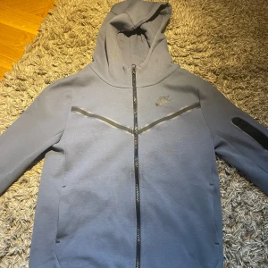 Ljusblå Nike tech från Nike - Säljer en Ljusblå tech fleece från Nike med dragkedja och huva. Tröjan har en stilren design med diskret Nike-logga på bröstet och en praktisk ficka på ärmen. Perfekt för en avslappnad stil. I storlek 147-158 cm
