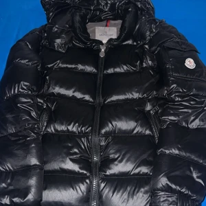 Svart dunjacka från Moncler - Säljer en snygg svart dunjacka från Moncler med dragkedja och huva. Jackan har ett stilrent quiltat mönster och Moncler-logga på ärmen. Perfekt för kalla dagar.