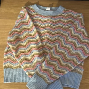 Mönstrad stickad tröja från Lindex - Säljer en färgglad stickad tröja från Lindex med ett zigzag-mönster i blått, rosa och beige. Tröjan har långa ärmar och ribbade muddar vid ärmslut och hals. Perfekt för att liva upp garderoben!