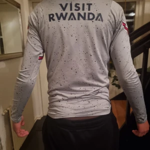Grå långärmad fotbollströja från Jordan x PSG - Snygg grå långärmad fotbollströja med svart och rött mönster. Tröjan har texten 'Visit Rwanda' på ryggen och 'Accor Live Limitless' på framsidan. Perfekt för fotbollsfans som gillar stil och komfort.