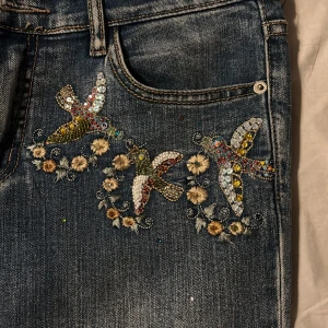 Blå jeans med broderade detaljer - Snygga blå bootcut jeans med unika broderade blommor och fåglar på framsidan. De har en klassisk femficksdesign och en bekväm passform. Säljer då dem tyvärr var för stora på mig.