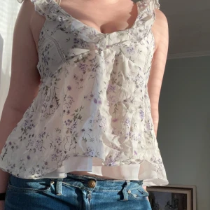 Blommig topp med volangdetaljer - Säljer en söt blommig topp i vitt med lila och gröna blommor. Toppen har volangdetaljer och öppen rygg. Perfekt för sommaren🌸