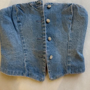 Blå jeanskorsett med knappar - Trendig blå jeanskorsett med dekorativa knappar framtill. Perfekt för att ge en edgy touch till din outfit. Korsetten har en strukturerad passform och är tillverkad i ett klassiskt denimtyg.