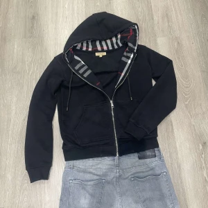 Svart hoodie från Burberry - Snygg svart hoodie från Burberry med dragkedja och randigt foder i huvan. Perfekt för en stilren och bekväm look. Den har långa ärmar och en klassisk passform.
