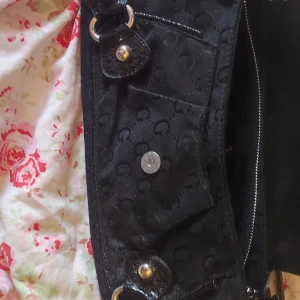 Svart axelväska med mönster och detaljer - Snygg svart axelväska med ett diskret mönster och glänsande detaljer. Väskan har en stor G-logga framtill och flera metallspännen. Den har en dragkedja för säker förvaring och en justerbar axelrem. Perfekt för att ge en stilren touch till din outfit.