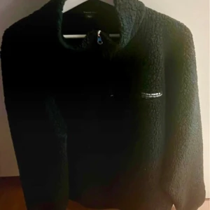 Svart fleece tröja från Urban Classics - Mysig svart fleece tröja från Urban Classics med halv dragkedja framtill. Perfekt för kyliga dagar och har en enkel design med en liten logga på bröstet.