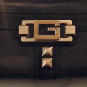 Svart plånbok från Guess - Snygg svart plånbok från Guess med gulddetaljer och en stor G-logga på framsidan. Plånboken har en dragkedja upptill och dekorativa nitar. Perfekt för att ge en elegant touch till din outfit.