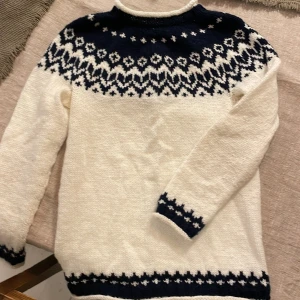 Vit stickad tröja med mönster - Säljer en vit stickad tröja med ett vackert mörkblått mönster runt halsen och ärmsluten. Tröjan har långa ärmar och är perfekt för kyliga dagar. Passar bra till både vardag och speciella tillfällen. Obs! Stl XXS