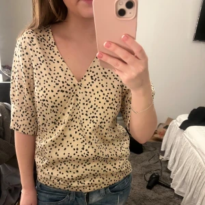 Beige prickig omlottopp - Säljer en snygg beige omlottopp med svarta prickar. Toppen har korta ärmar och en v-ringad design som ger en stilren look. Perfekt för både vardag och speciella tillfällen.