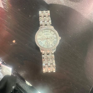 Silverfärgad klocka från Geneva - Elegant silverfärgad klocka från Geneva med glittrande detaljer runt urtavlan och länken. Klockan har en klassisk design med romerska siffror och tre mindre urtavlor inuti. Perfekt för att ge en touch av glamour till din stil.