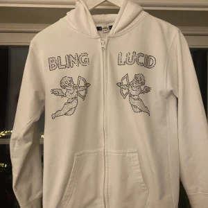 Vit hoodie från Bling Lucid - Säljer en vit hoodie från Bling Lucid med glittrande text och änglamotiv på framsidan. Hoodien har dragkedja och är tillverkad i en bomulls- och polyesterblandning. Tyvärr har några glitterstenar ramlat av (se bild 3 och 4)..