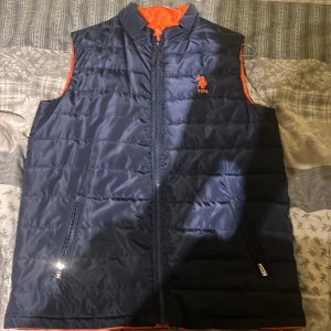 Vändbar dunväst från USPA - Säljer en snygg vändbar dunväst från USPA. Ena sidan är marinblå med orange broderad logga, och den andra sidan är orange med svart broderad logga. Västen har dragkedja och två fickor med dragkedjor på båda sidor.