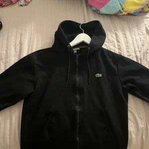 Svart hoodie från Lacoste - Säljer en svart hoodie från Lacoste med dragkedja och huva. Den har en klassisk passform och det ikoniska krokodilmärket på bröstet. Perfekt för en avslappnad stil.