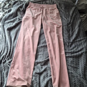 Rosa mjukisbyxor från Juicy Couture - Snygga rosa mjukisbyxor från Juicy Couture med dragsko i midjan och praktiska fickor både fram och bak. Byxorna har en broderad logga på framsidan och är perfekta för en avslappnad stil. Tillverkade i mjukt material för extra komfort. 