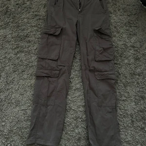 Grå cargobyxor från Bershka - Säljer ett par grå cargobyxor från Bershka med flera praktiska fickor och dragkedja. Perfekta för en avslappnad stil med en loose passform.