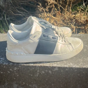 Valentino sneakers med svart detalj - Snygga Valentino sneakers i vitt med en bred svart rem över snörningen. Skorna har en unik design med nitar runt hälen och en klassisk siluett. Perfekta för en stilren look.