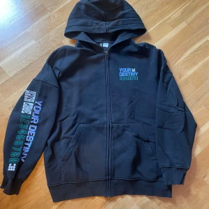 Svart hoodie från H&M Divided - Snygg svart hoodie från H&M Divided med tryck 'Your Destiny' i blått och lila på både framsidan och baksidan. Den har en dragkedja framtill och en luva. Perfekt för en cool och avslappnad stil.