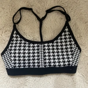 Svart och vit sporttopp från Nike - Säljer en snygg sporttopp från Nike i svart och vitt med ett klassiskt hundtandsmönster. Toppen har justerbara axelband och är tillverkad i Dri-FIT-material för optimal komfort under träning. Hör av er vid frågor❤️