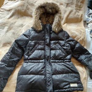 Svart jacka från Odd Molly - Svart jacka/puffer jacket från Odd Molly med pälskläddhuva (även insida). Jackan har både dragkedja och knappar framtill för, spänne för att dra åt i midjan och snören nedtill. Perfekt för kalla vinterdagar med sin varma och vadderade design. Finns även en ficka på insidan.