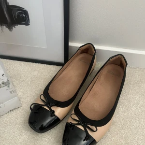 Ballerinaskor - Stilrena ballerinaskor i beige och svart med en elegant rosett på tån. Skorna har en rundad tå och en låg klack, vilket gör dem bekväma för daglig användning. Perfekta för en chic och avslappnad look.