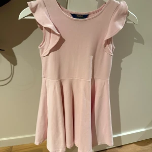 Rosa klänning från Ralph Lauren - Söt rosa klänning från Ralph Lauren med volangdetaljer vid ärmarna. Klänningen har en rund hals och är ärmlös, vilket gör den perfekt för varmare dagar. Den har en fin, fladdrig kjol som ger en lekfull känsla. Barnstorlek 8-10. Liten i storlek. 