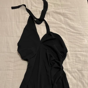 Svart långklänning med knytband - Helt ny snygg svart klänning med halterneck-design och draperad detalj framtill. Klänningen har en slits baktill som ger extra rörelsefrihet. Perfekt för en somrig look! Midiklänning! Storlek M. 