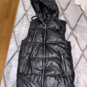 Svart dunväst med huva - Snygg svart dunväst från Zara med dragkedja och huva. Perfekt för kyliga dagar när du vill hålla stilen. Västen har praktiska fickor med dragkedjor.