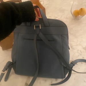 Blå ryggsäck från Michael Kors - Snygg blå ryggsäck från Michael Kors i skinn. Väskan har en praktisk dragkedja framtill och justerbara axelremmar. Insidan är rymlig med flera fack för enkel organisering. Perfekt för både vardag och utflykter.