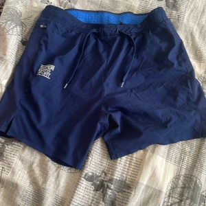 Mörkblå shorts från Superdry - Snygga mörkblå shorts från Superdry med snörning i midjan. Perfekta för en avslappnad stil. De har en liten logga på sidan och är gjorda i ett lätt material som passar bra för varma dagar.