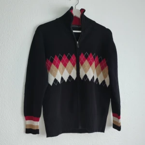 Svart tröja med argylemönster från Pahladium - Säljer en svart tröja från Pahladium med ett klassiskt argylemönster i rött, beige och vitt. Tröjan har en dragkedja framtill och ribbade muddar vid ärmslut och nederkant. Perfekt för en stilren look!