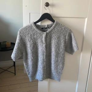 Grå kofta från Gina Tricot - Mysig grå kofta från Gina Tricot med korta ärmar och knappar framtill. Perfekt för en stilren och bekväm look. Den har en fluffig textur som ger extra värme och komfort.