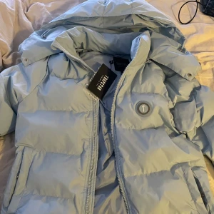 Ljusblå pufferjacka från Trapstar - Säljer en ljusblå pufferjacka från Trapstar med huva och dragkedja. Jackan har en stor logga på ryggen och är perfekt för kyliga dagar. Den är både stilren och funktionell.