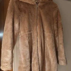 Beige pälsjacka med huva, jakke - Säljer en mysig beige pälsjacka med huva och dragkedja framtill. Jackan har långa ärmar och är perfekt för kyligare dagar. Den är både stilren och bekväm, vilket gör den till ett måste i garderoben.