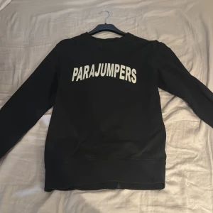 Svart sweatshirt från Parajumpers - Säljer en svart sweatshirt från Parajumpers med vit text på framsidan. Tröjan har långa ärmar och en patch på ärmen. Perfekt för en stilren och bekväm look.