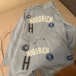 Ljusblå hoodie och byxor från Hoodrich - Säljer ett set med ljusblå hoodie och matchande byxor från Hoodrich. Hoodien har en stor logotyp framtill och flera märken i blått och vitt. Byxorna har samma design med logotyp och märken. Perfekt för en avslappnad stil.