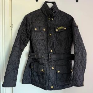 Svart quiltad jacka från Barbour - Snygg svart quiltad jacka från Barbour med bälte i midjan och guldiga knappar. Jackan har flera praktiska fickor och en hög krage med spänne. Perfekt för kyligare dagar med sin stilrena design och funktionella detaljer.