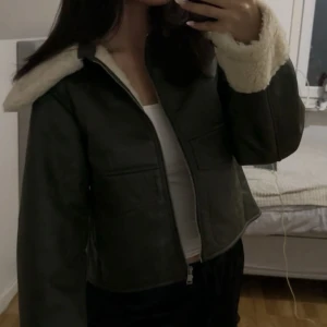 Grön jacka med fuskpäls - Snygg grön jacka med vit fuskpäls på kragen och ärmsluten. Jackan har en dragkedja framtill och är croppad i modellen. Perfekt för kyligare dagar när du vill vara både varm och stilren.