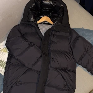 Svart dunjacka från Moncler - Säljer en snygg svart dunjacka från Moncler. Jackan har en pufferdesign med långa ärmar och en praktisk huva. Den är utrustad med dragkedja och knappar för extra skydd mot kyla. Perfekt för kalla vinterdagar.  Storlek L, men passar M