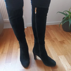 Svarta höga mocka stövlar  med dragkedja - Snygga svarta stövlar i mocka med hög klack och dragkedja på sidan. Perfekta för en stilren look under höst och vinter. De når över knäet och har en elegant design  med äkta päls inuti