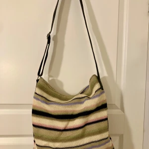 Randig axelväska i stickat material (Ceannis) - Snygg axelväska från Ceannis med ett färgglatt randigt mönster i beige, svart, grön, rosa och blå. Väskan har en praktisk dragkedja och är tillverkad i ett stickat material. Storlek: 40x35