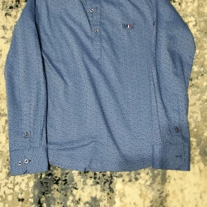 Blå pikétröja med mönster - Snygg blå pikétröja med diskret mönster och knappar framtill. Tröjan har långa ärmar och en liten broderad logga på bröstet. Perfekt för en stilren look.