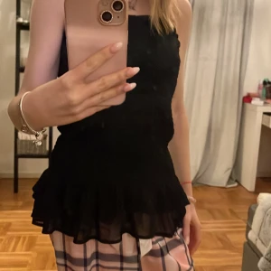 Svart axelbandslös topp - Snygg svart axelbandslös topp med volangdetaljer i nederkant. Perfekt för en stilren look. Toppen har en tight passform som framhäver figuren.