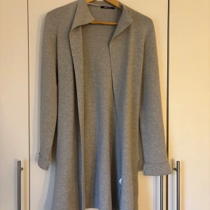 Grå lång kofta från Gina Tricot - Säljer en grå lång kofta från Gina Tricot. Koftan har långa ärmar och ett knytband i midjan för en justerbar passform. Hängt i garderoben ett tag och inte kommit till användning, finns inga fläckar eller liknande utan är i ett väldigt bra skick. jackan/koftan är i storlek xs men passar även M/S